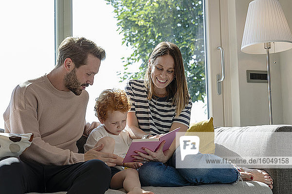Lächelnde Frau  die ein Bilderbuch liest  während sie neben ihrem Sohn und einem Mann auf dem Sofa im heimischen Wohnzimmer sitzt