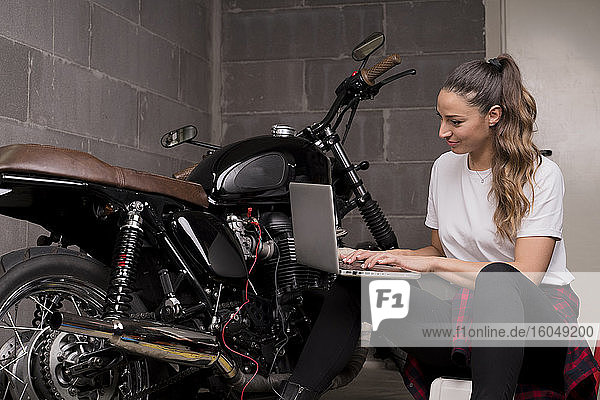 Junge Frau  die einen Laptop benutzt und neben einem Motorrad sitzt