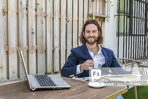 Lächelnder gutaussehender junger Geschäftsmann liest Zeitung und hält eine Kaffeetasse neben seinem Laptop am Tisch in einem Café