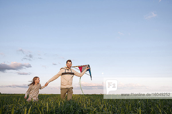 Fröhlicher Vater und Tochter mit Drachen auf grüner Landschaft gegen den Himmel laufend