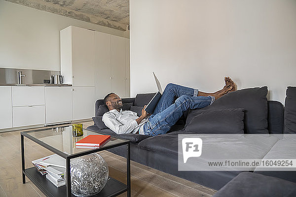 Lächelnder Mann  der in einer modernen Wohnung auf der Couch liegt und einen Laptop benutzt