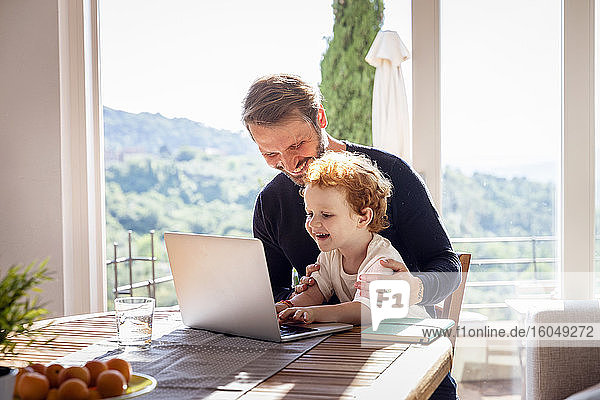 Smiling man looking at son using laptop while sitting against window in living room