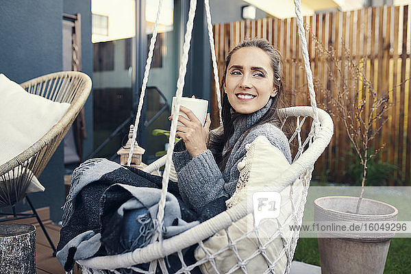 Lächelnde junge Frau hält Tasse  während sie sich auf einer Schaukel im Garten entspannt