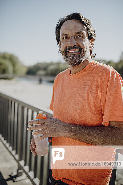 Portrait of smiling man holding water bottle while standing against clear sky in park