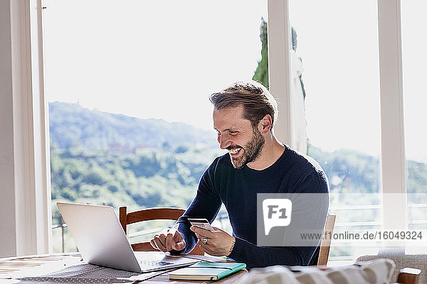 Lächelnder gut aussehender Mann  der eine Kreditkarte hält  während er einen Laptop am Esstisch vor dem Fenster benutzt