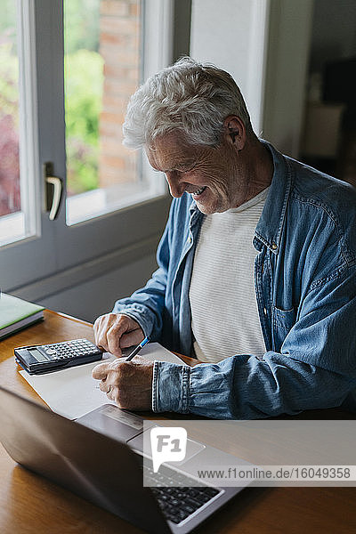 Glücklicher älterer Mann  der zu Hause mit Taschenrechner und Laptop auf Papier schreibt