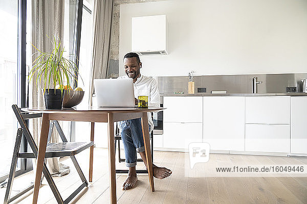 Porträt eines lächelnden Mannes  der an einem Tisch in einer modernen Wohnung sitzt und einen Laptop und Kopfhörer benutzt