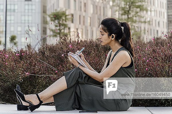 Weibliche Fachkraft schreibt in einen Notizblock  während sie auf einem Sitz bei Pflanzen sitzt