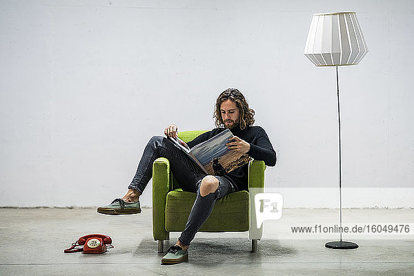 Young man reading book while relaxing on armchair against wall at home