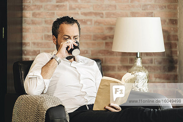 Mature man sitting in armchair  reading book  drinking coffee