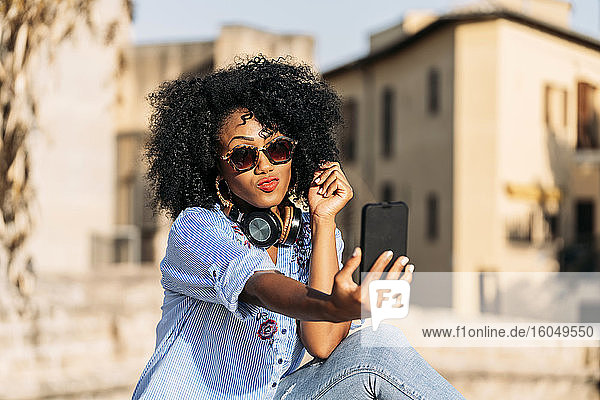Lächelnde Frau mit Afro-Haar und Sonnenbrille macht ein Selfie mit ihrem Smartphone
