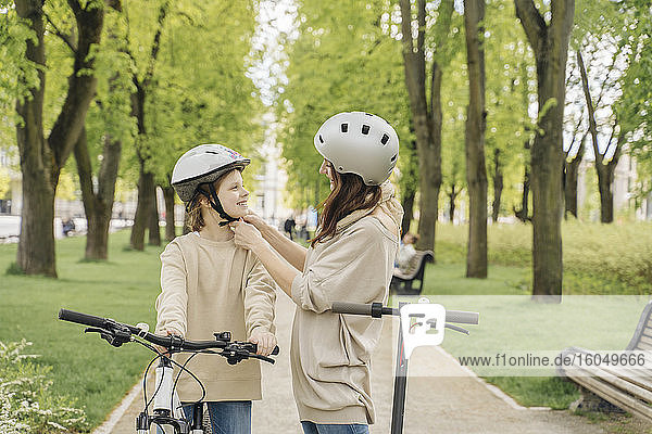 Mutter  die ihrer Tochter einen Helm trägt  während sie im Stadtpark vor Bäumen steht