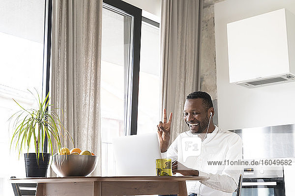 Porträt eines glücklichen Mannes  der während eines Videochats am Küchentisch sitzt und ein Siegeszeichen zeigt