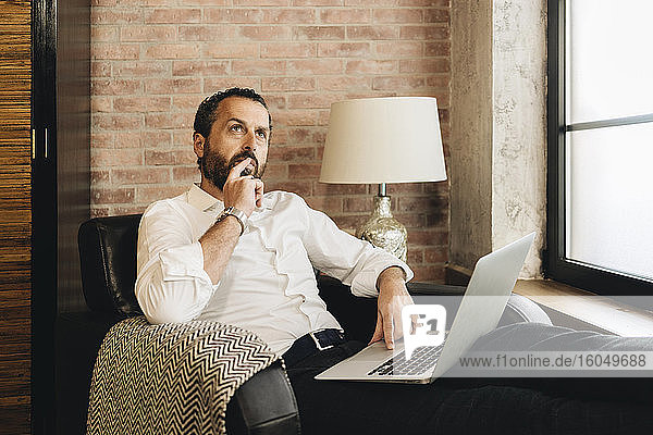 Mature man sitting in armchair  using laptop  thinking