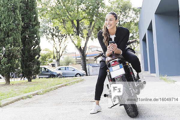 Lächelnde Frau  die auf einem Motorrad sitzt und ein Smartphone benutzt