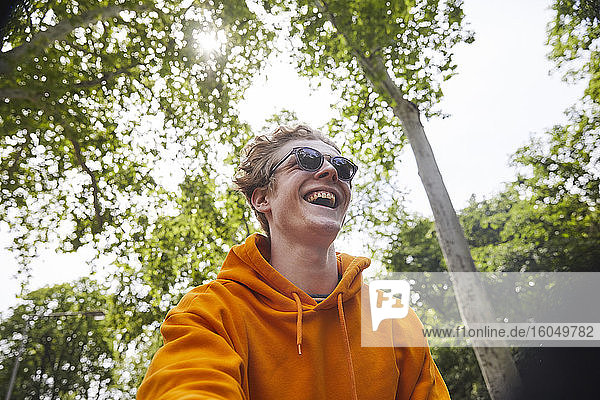 Porträt eines lachenden jungen Mannes mit Sonnenbrille und orangefarbenem Kapuzenshirt in der Natur
