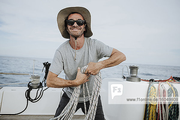 Lächelnder Seemann mit Sonnenbrille und Hut  der Taue in einem Segelboot hält