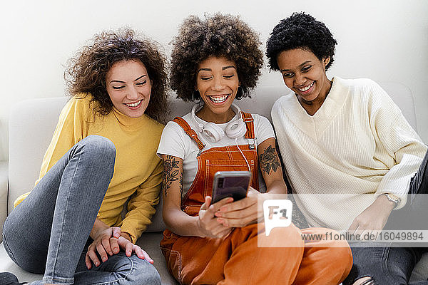 Gruppenbild von drei lachenden Freunden  die auf der Couch sitzen und ein Selfie mit ihrem Smartphone machen