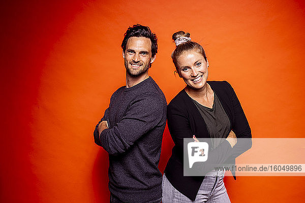 Lächelndes erwachsenes Paar  das mit verschränkten Armen vor einem orangefarbenen Hintergrund steht