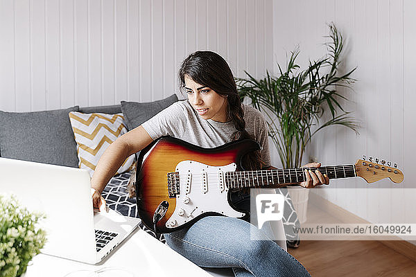 Junge Frau benutzt einen Laptop  während sie zu Hause E-Gitarre spielt
