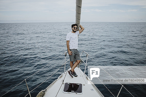 Male sailor wearing sunglasses standing on bow of sailboat in sea