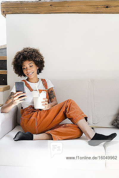 Porträt einer lächelnden jungen Frau  die zu Hause auf der Couch sitzt und eine Schachtel mit asiatischem Essen auf ihr Handy schaut