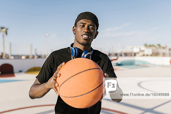 Junger Mann hält Basketball  während er an einem sonnigen Tag vor einem klaren Himmel steht