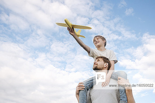 Mann  der seinen Sohn auf den Schultern trägt und mit einem Spielzeugflugzeug gegen den Himmel spielt