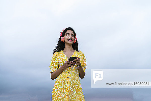 Nachdenkliche Frau hält ihr Smartphone in der Hand und hört Musik gegen den Himmel