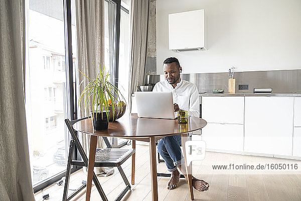 Porträt eines Mannes  der in einer modernen Wohnung am Tisch sitzt und einen Laptop und Kopfhörer benutzt