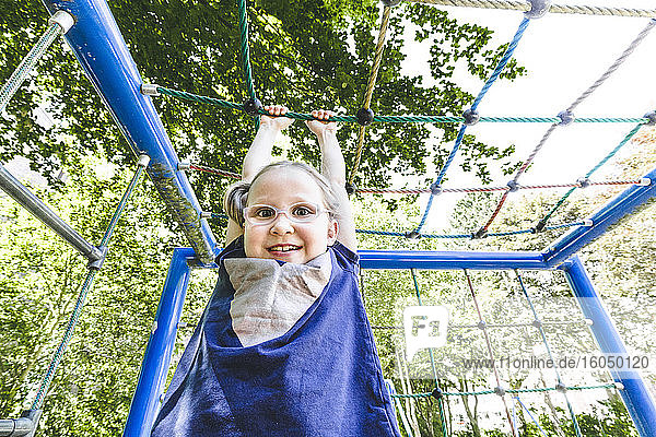 Lächelndes süßes Mädchen hängt am Klettergerüst auf einem Spielplatz im Park