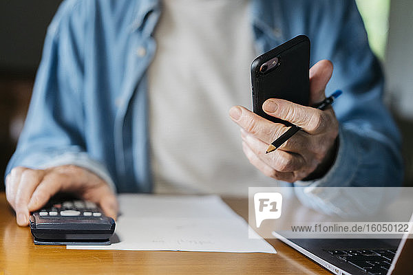 Ein älterer Mann hält ein Smartphone in der Hand  während er zu Hause einen Taschenrechner und einen Laptop benutzt