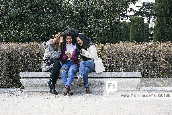 Lächelnde Freundinnen benutzen ihr Smartphone  während sie auf einer Parkbank vor Pflanzen sitzen