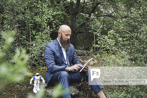 Bald businessman using digital tablet while sitting with robot amidst trees in forest