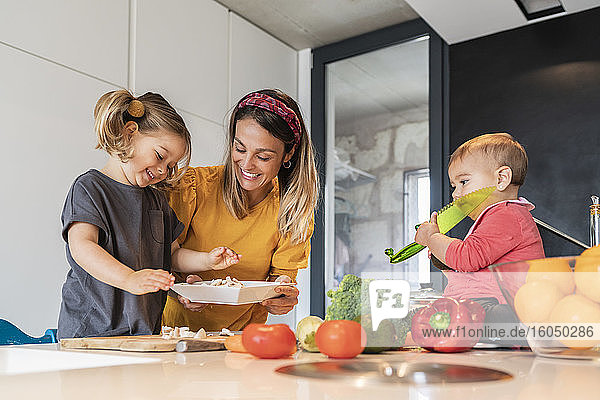 Lächelnde Mutter und Mädchen bereiten Essen zu  während die kleine Tochter auf der Kücheninsel sitzt