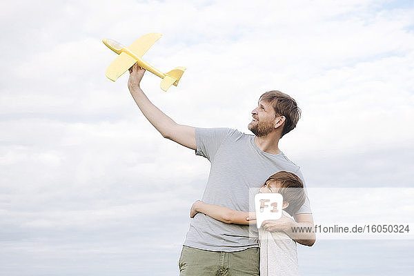 Glücklicher Junge  der seinen Vater umarmt und ein Spielzeugflugzeug hält  das gegen den Himmel steht