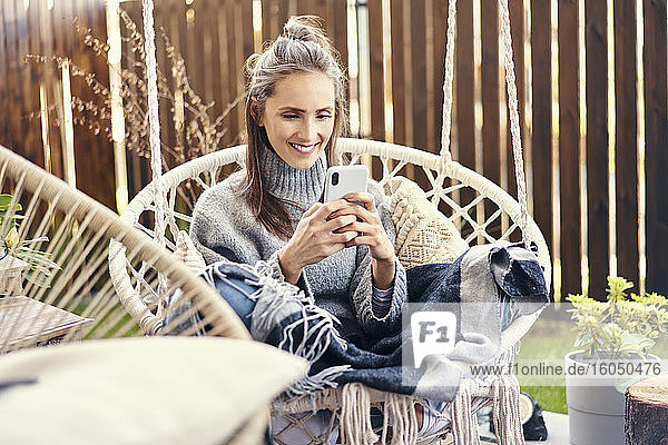Lächelnde junge Frau benutzt ihr Smartphone  während sie sich auf einer Schaukel im Garten entspannt
