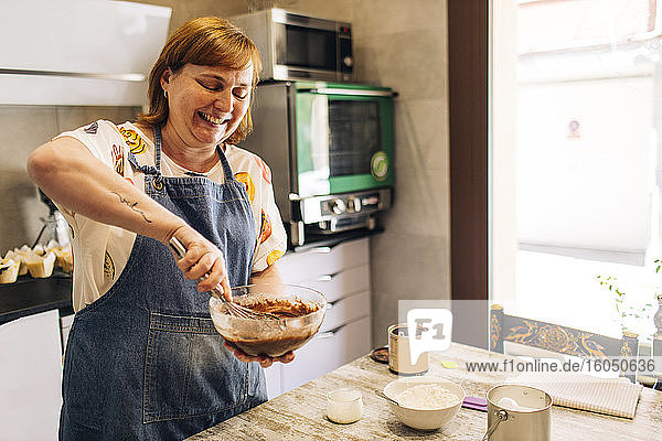 Lächelnde Bäckerin mischt Kuchenteig in einer Schüssel auf einem Tisch in einer Werkstatt
