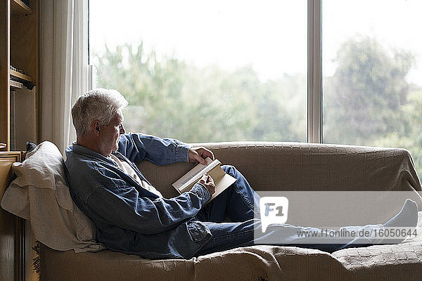 Älterer Mann liest ein Buch und entspannt sich auf dem Sofa am Fenster