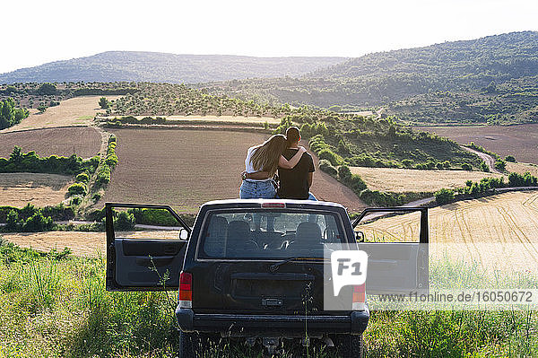 Mid adult couple sitting on 4x4 vehicle roof while looking at landscape during road trip