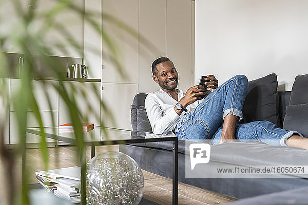 Porträt eines lächelnden Mannes  der auf einer Couch in einer modernen Wohnung sitzt und ein Smartphone benutzt