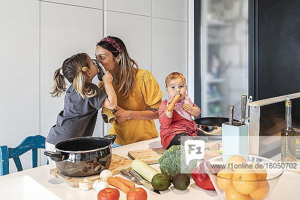 Mutter und Tochter küssen sich durch den Deckel  während das kleine Mädchen auf der Kücheninsel zu Hause sitzt