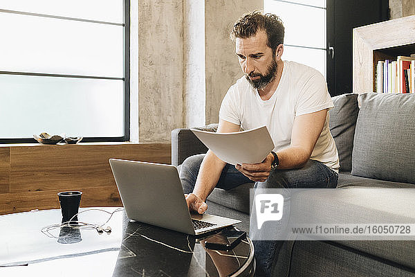 Mature man sitting on couch  using laptop  holding papers