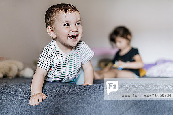 Porträt eines glücklichen kleinen Mädchens  das auf dem Bett krabbelt  während ihre Schwester im Hintergrund sitzt