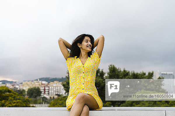 Entspannte junge Frau sitzt auf einer Stützmauer auf einer Terrasse gegen den Himmel