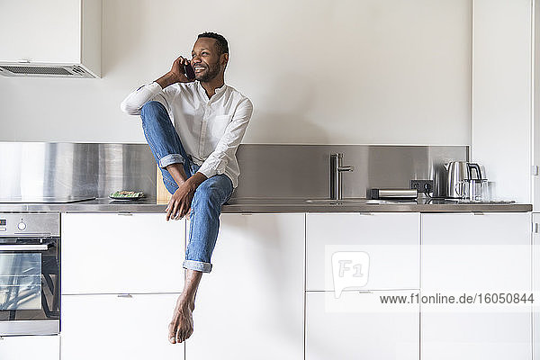 Portrait of smiling man on the phone sitting on kitchen counter at home looking at distance