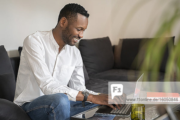 Lächelnder Mann sitzt zu Hause auf der Couch und benutzt einen Laptop