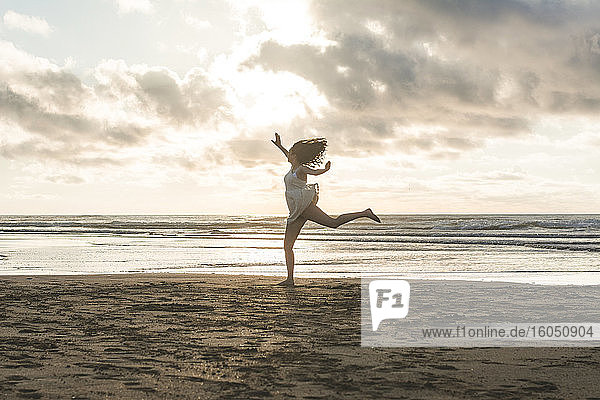 Unbeschwerte junge Frau springt am Strand gegen den bewölkten Himmel bei Sonnenuntergang