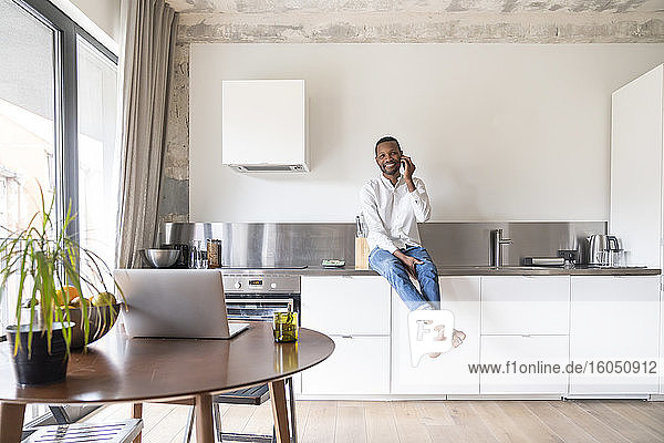 Porträt eines lächelnden Mannes am Telefon  der auf dem Küchentisch in einer modernen Wohnung sitzt