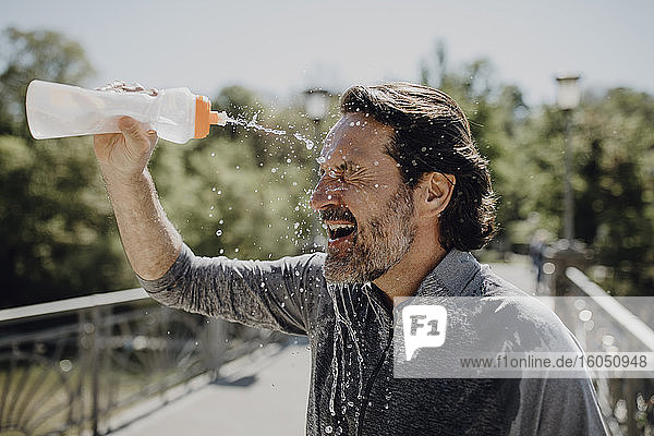 Nahaufnahme eines reifen Mannes  der sich Wasser ins Gesicht gießt  während er an einem sonnigen Tag im Park steht